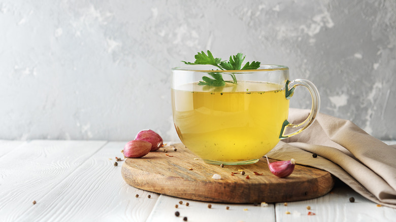 A glass cup of broth with a parsley leaf in it, surrounded by garlic cloves on a wooden plank