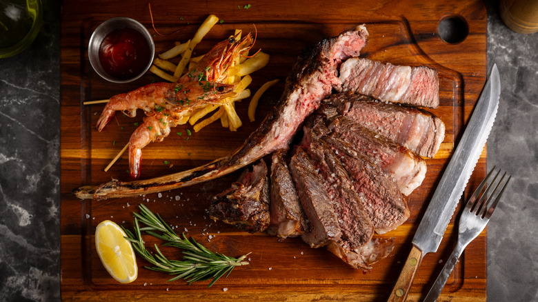 tomahawk steak next to shrimp on a wooden cutting board with a serrated knife and a fork