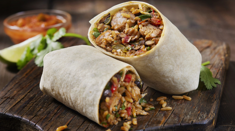 halved burrito with rice, meat, and beans on a wooden board with a lime wedge, cilantro, and salsa