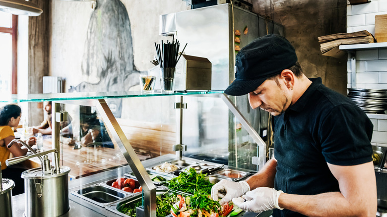 A worker builds a meal with vegetables behind the counter at a restaurant