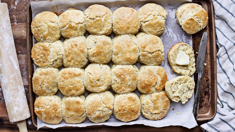 Pan of cooked Southern biscuits, one cut in half with a pat of butter, with a butter knife on one side and a flour-covered rolling pin on the other