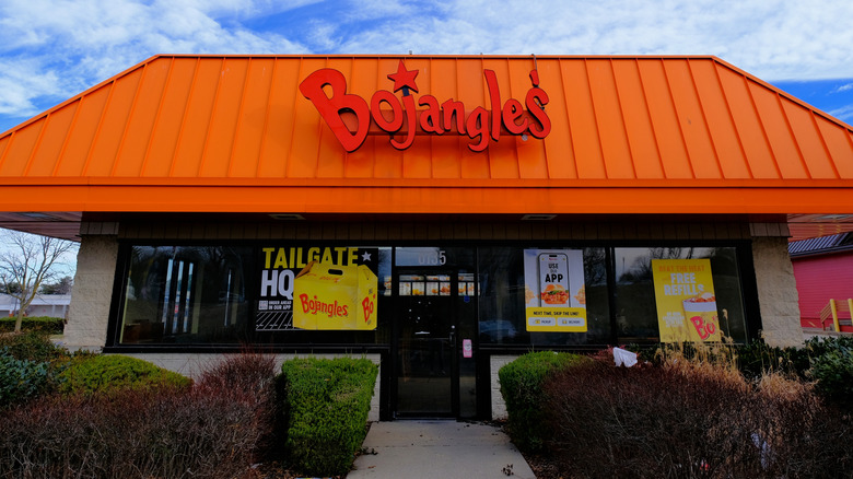 Front of a Bojangles restaurant with an orange facade with the name in red letters, bushes along the walkway to the door