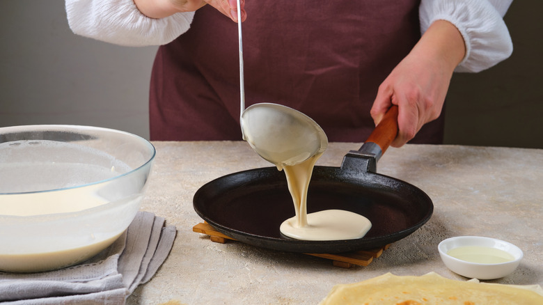 A person making crepes with a ladle in a shallow cast iron pan