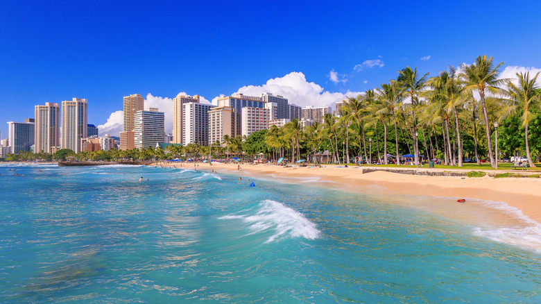 Tropical skyline with buildings, palm trees, sand beach, and aquamarine water against a blue sky