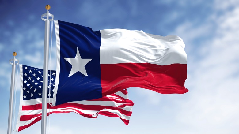 Red, white and blue Texas state star flag flying in front of U.S. flag against a sky with scattered clouds