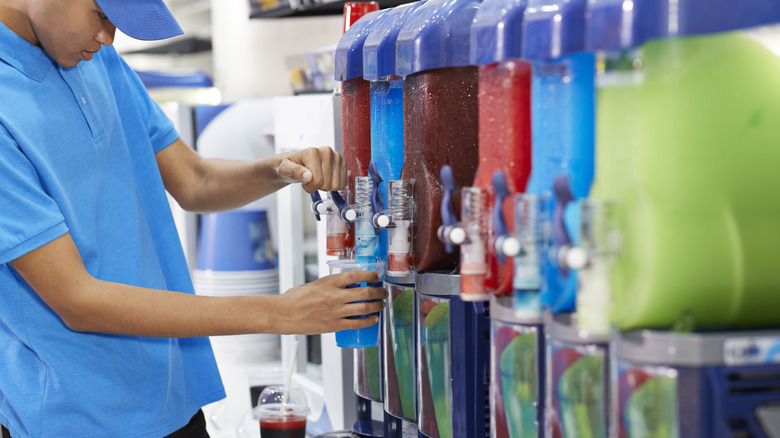 A young man in a blue baseball cap fills a cup from a slushie dispenser