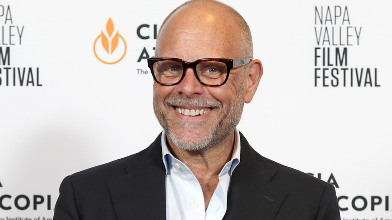 Alton Brown wears glasses and smiles on an event red carpet