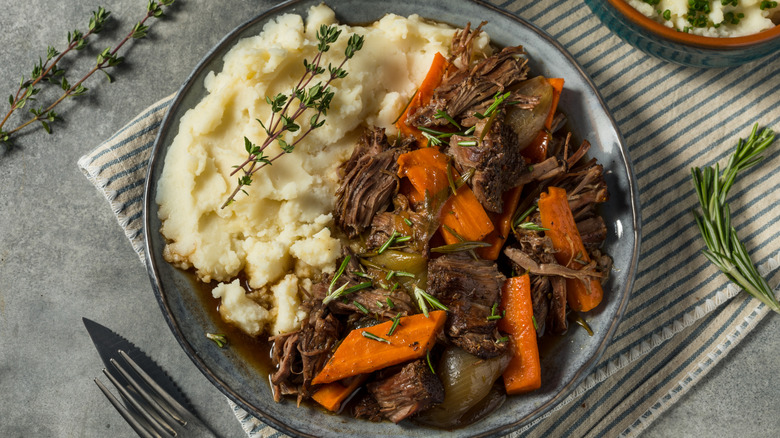 Pot roast on a plate with mashed potatoes, carrots, and herbs