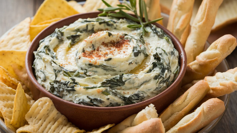 A bowl of spinach dip with chips and breadsticks, garnished with fresh rosemary