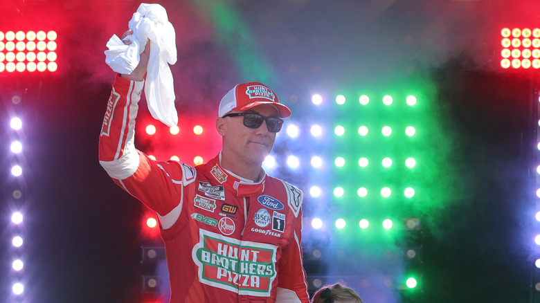 A NASCAR driver in a red Hunt Brothers Pizza racing suit waves to the crowd with red and green lights glowing in the background.