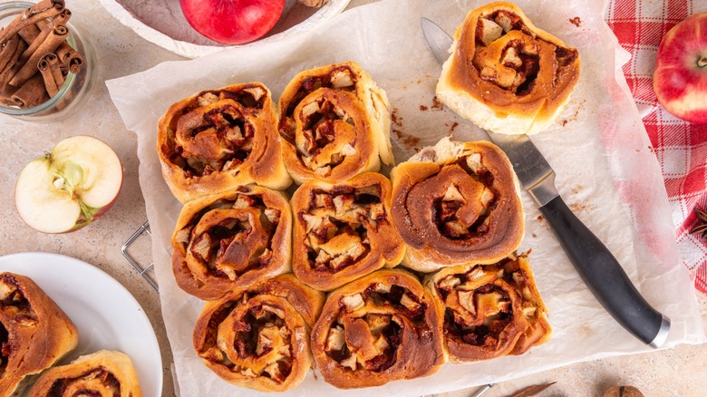 Apple pie cinnamon rolls on a piece of parchment paper, surrounded by apples and cinnamon sticks