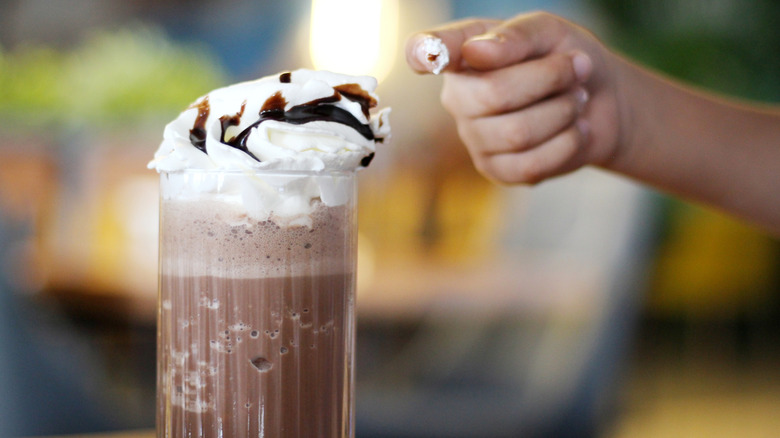 A hand taking a taste from a chocolate malt topped with whipped cream and chocolate syrup.