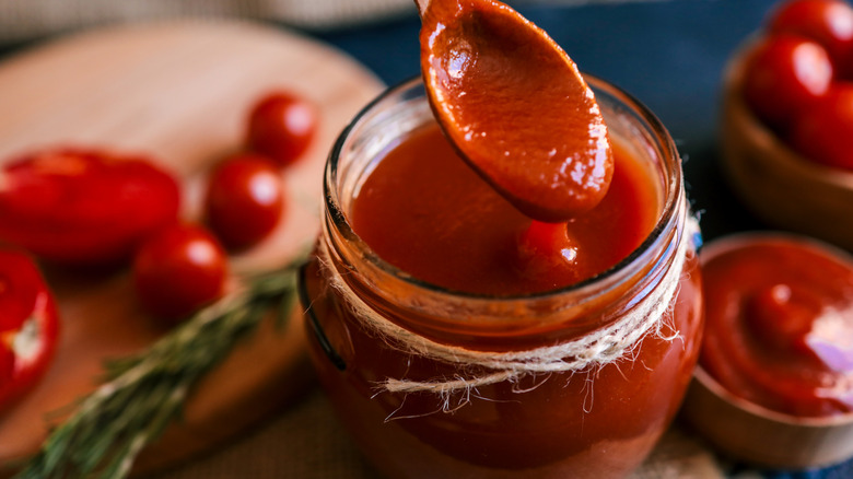 A spoonful of tomato sauce above a jar with a tomato and rosemary-filled cutting board in the background
