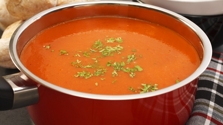 A red saucepan full of home made tomato soup, with parsley garnish, on a checkered tablecloth, and ready to serve.