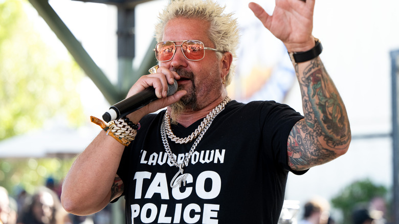 Guy Fieri speaking into a microphone at an event while wearing a shirt that says 