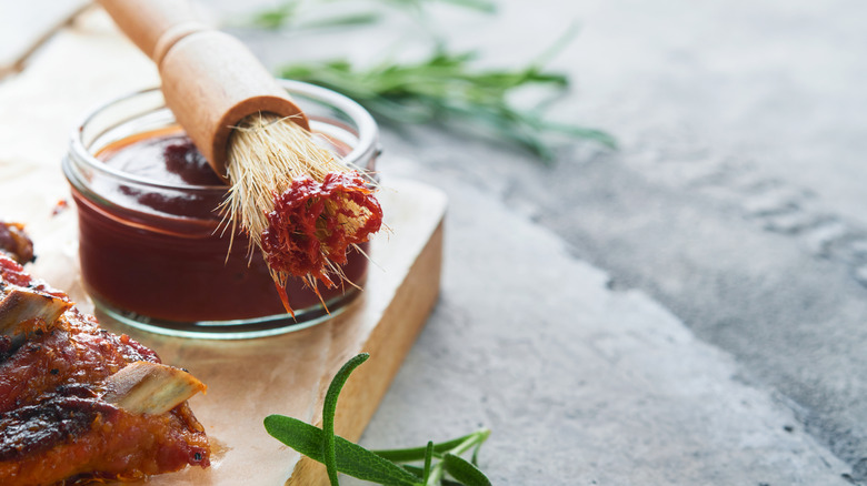 a barbecue brush sits on top of a dish of barbecue sauce, next to a close-up of barbecue ribs