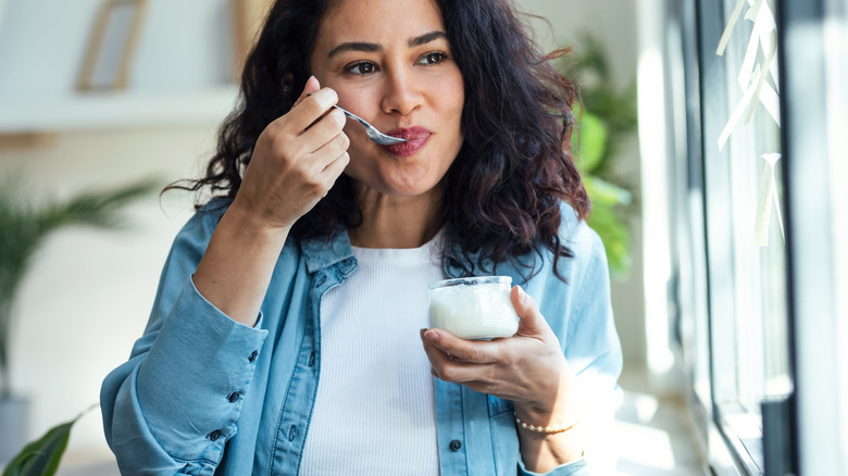 A woman stands by a window and eats yogurt from a small cup