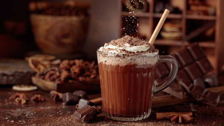 A cup of hot chocolate that's surrounded by pieces of chocolate and being topped with whipped cream and more chocolate