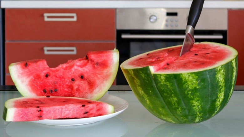 a halved watermelon with a knife in it and two watermelon slices on a plate sit on a counter