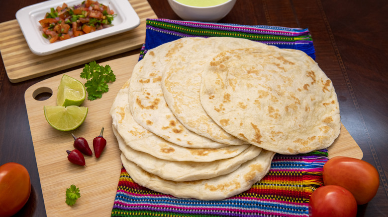 Large tortillas spread out in a fan on a colorful cloth, surrounded by tomatoes, lime pieces, peppers, and herbs