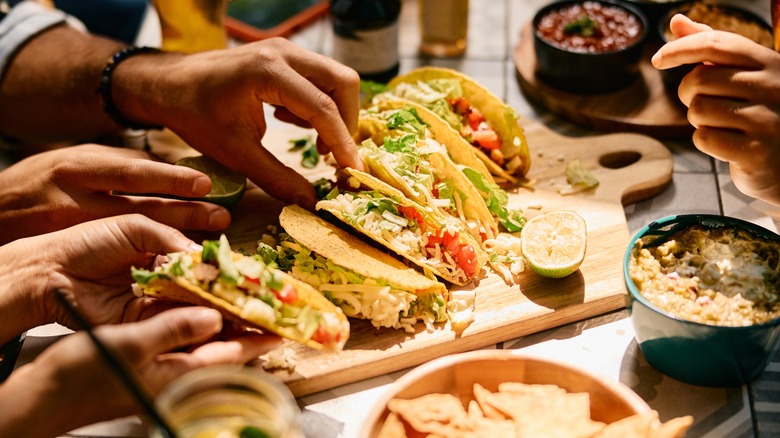 Close up of hands grabbing tacos off a wooden cutting board on a restaurant table