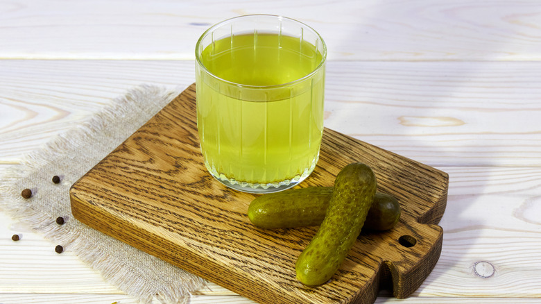 A glass of light green liquid with pickles on a wood board