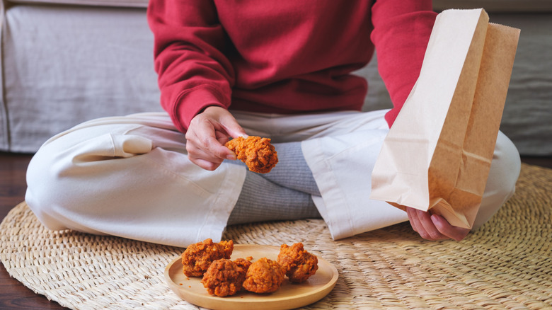 A person sitting cross-legged, holding fried chicken and a paper bag