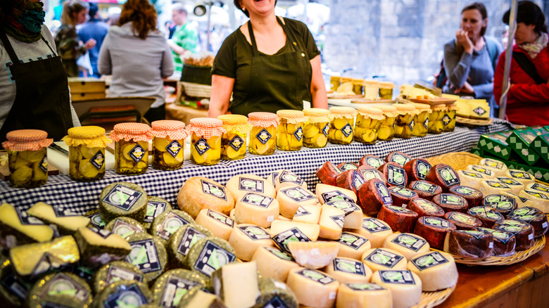 Preserves and cheese at a farmers market with vendors and shoppers in the background
