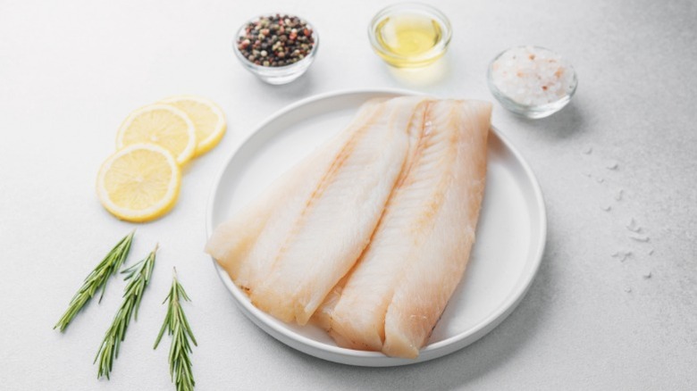 Raw pollock fillet on a plate, surrounded with herbs and seasoning.