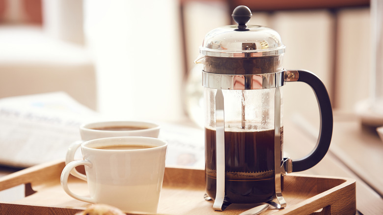 A French press half full of coffee on a breakfast tray next to two mugs.