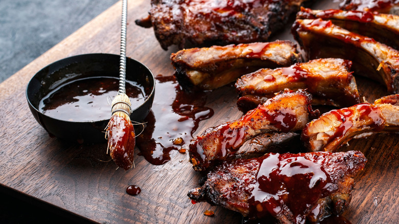 A cup of barbecue sauce next to ribs on a wooden board
