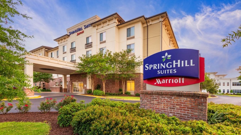 The exterior of a SpringHill Suites by Marriott with a blue and red sign and lots of greenery in front.