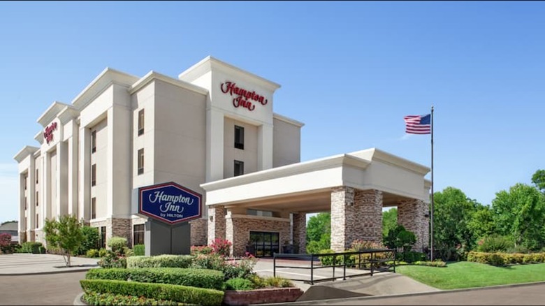 The exterior of a Hampton Inn with a blue sign and a flagpole out front.