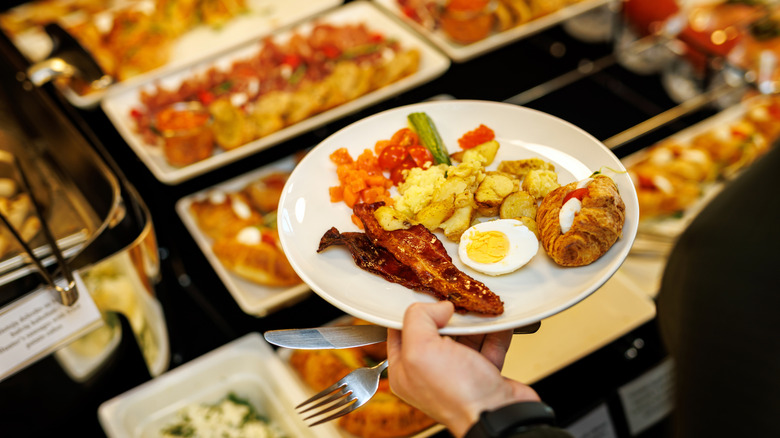 A person holds a plate with bacon, potatoes, a croissant, and a hard-boiled egg. There are several serving dishes full of breakfast food in background