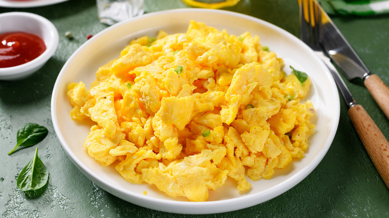 A white plate of scrambled eggs next to utensils and ketchup