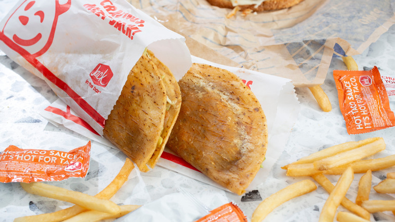 Two Jack in the Box tacos on a paper surface near French fries and hot sauce packets