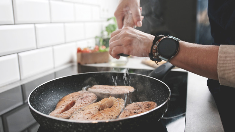A chef's hands adds seasoning to fish fillets in a pan on a stovetop