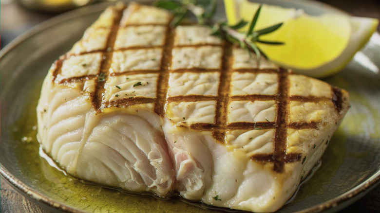 A large slab of grilled swordfish on a plate with a rosemary sprig and lemon slice