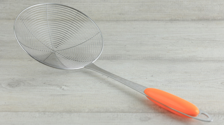 Stainless steel spider strainer with orange handle on a gray surface with wood finish.