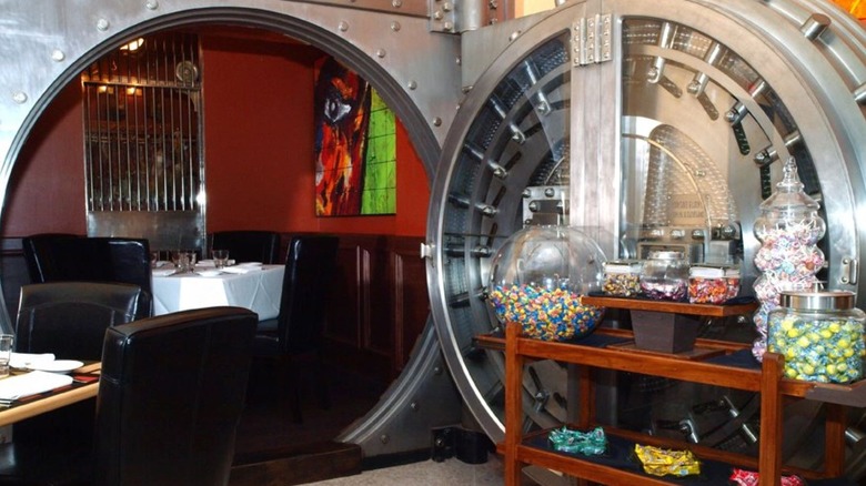 Open silver bank vault with a table and four chairs inside, part of another table outside and a wooden rack with bowls of candy