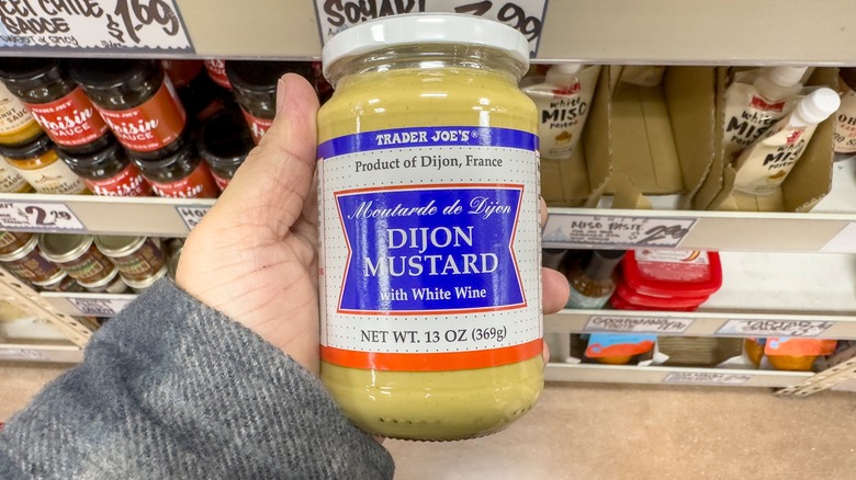 A hand holds a jar of Trader Joe's Dijon mustard in front of a shelf of assorted condiments