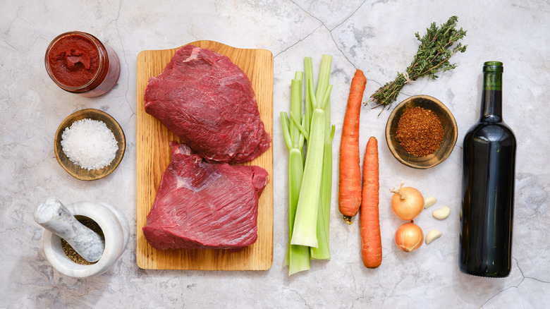 Ingredients for beef stew including raw beef, celery, onions, garlic, carrots, tomato paste, red wine, herbs, and spices