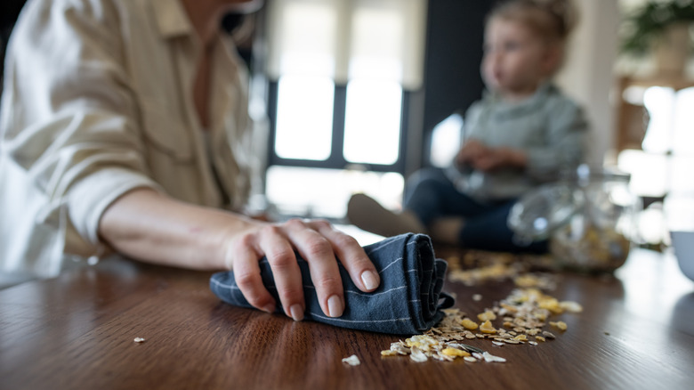 Person gathering scattered cereal on counter with towel with toddler blurred in background