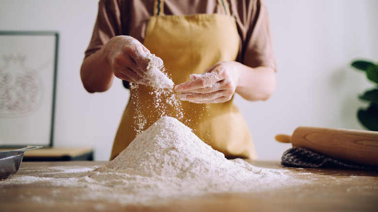 A woman in a yellow apron using her hand to pour flour onto a larger pile of flour.