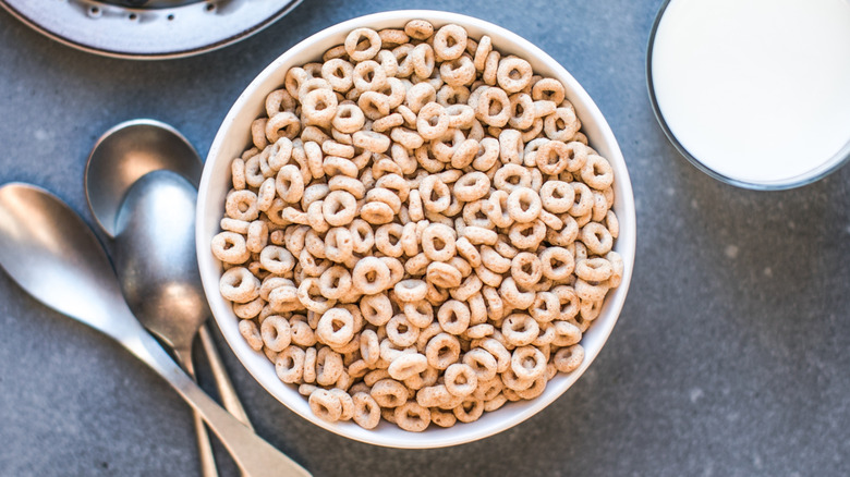 A bowl of Cheerios with a glass of milk and spoons on a cement surface