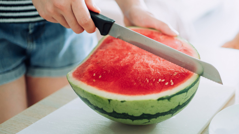 A person cutting half a watermelon with a bread knife