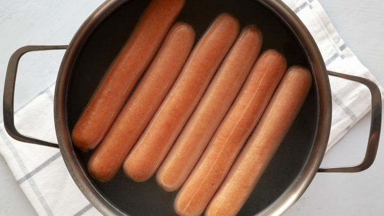A pot with hot dogs in water sitting on a dish towel on a white surface.