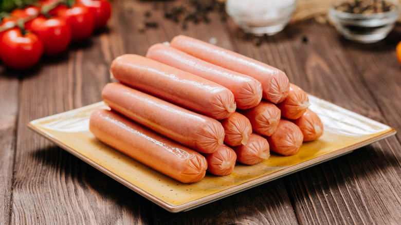 Stack of hot dogs on a plate with tomatoes on the vine, salt, and pepper in the background on a wood picnic table.
