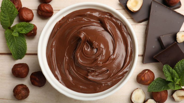 Chocolate hazelnut spread in bowl surrounded by chocolate, hazelnuts, and mint leaves