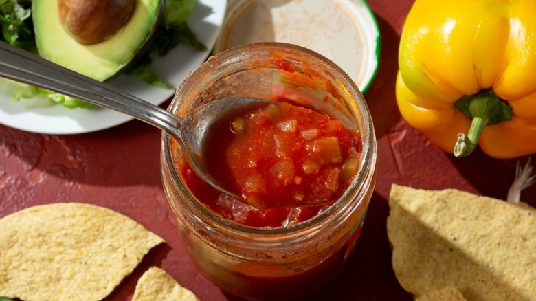 Top view of a spoon in a jar of salsa surrounded by chips, bell pepper, and avocado